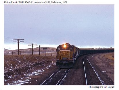 In this 1970s colour photograph taken by the railfan designer Ian Logan as he travelled across Nebraska in the cab of the California Zephyr. a train approaching from the west is headed by the EMD SD40-2 locomotive 3256, headlight blazing in the fading light, patches of snow lying next to the tracks, telegraph poles marching into the distance.
