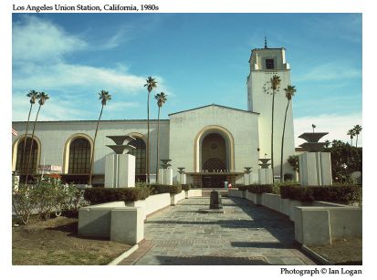 In this colour photograph taken by the railfan designer Ian Logan, Los Angeles Union Station stands proud with its clock tower at the end of a patterned broad walk and open space planted with tall thin palm trees. 