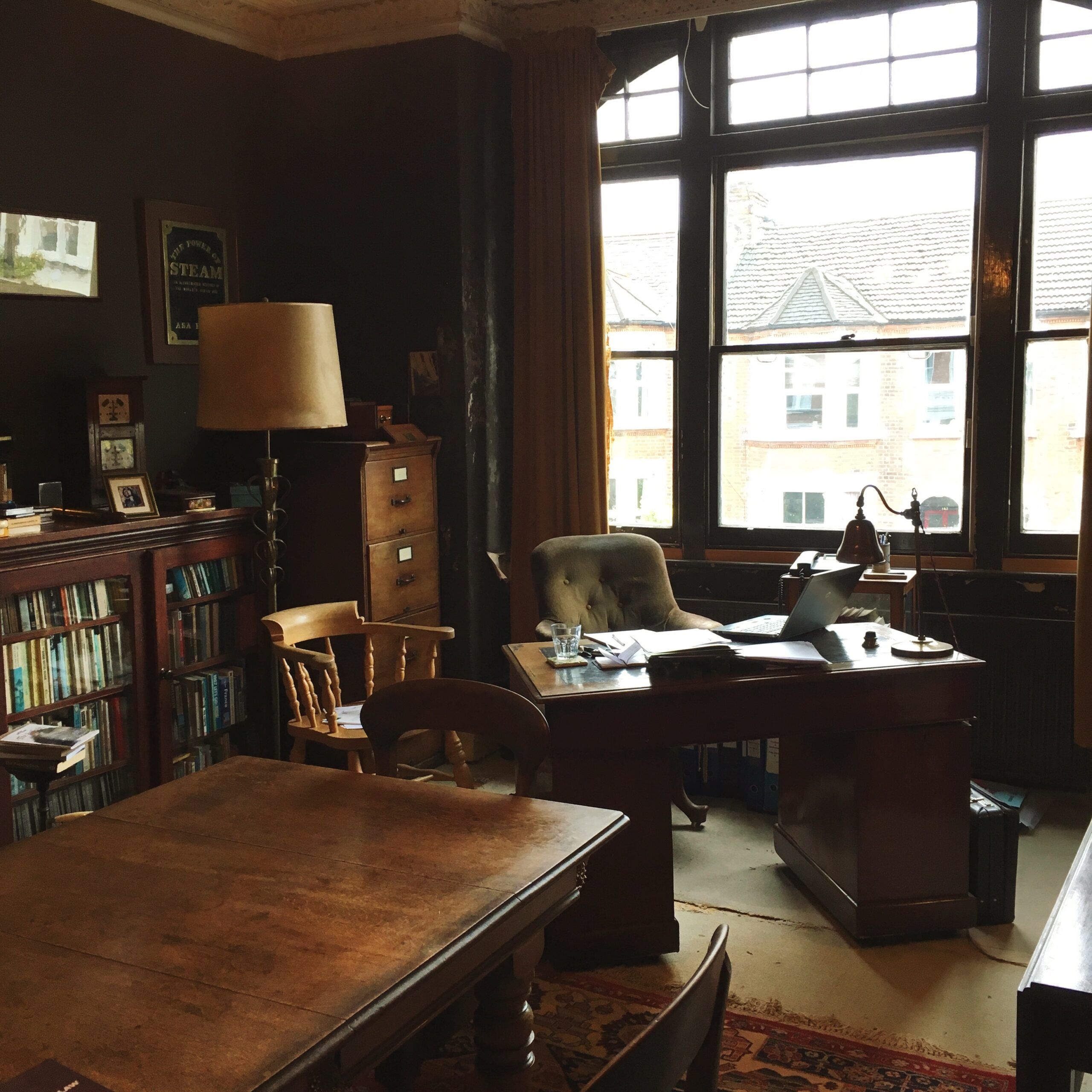 A wooden pedestal desk with a green armchair, in a study with a dark wooden able and lots of bookcases and file cabinets. 