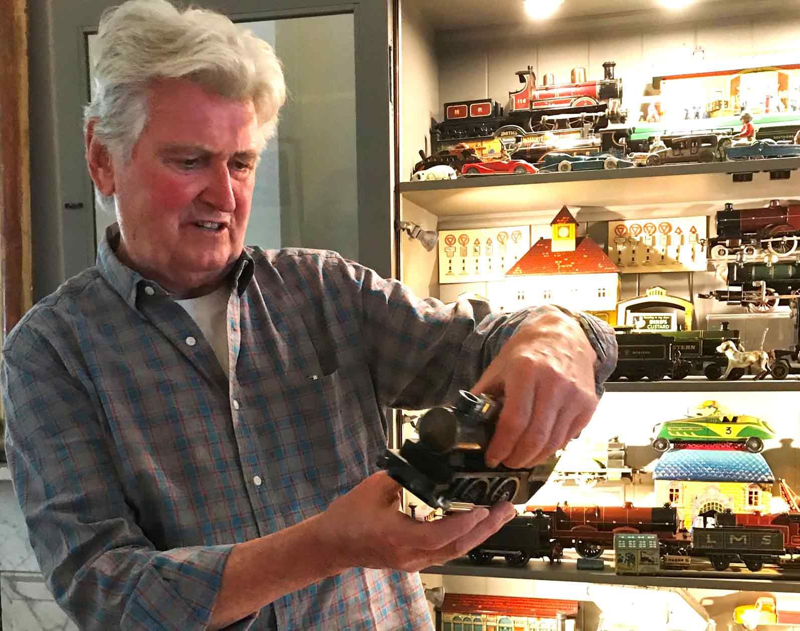 In this color photograph, the designer Ian Logan holds a model steam locomotive with an illuminated cabinet behind him containing other models.