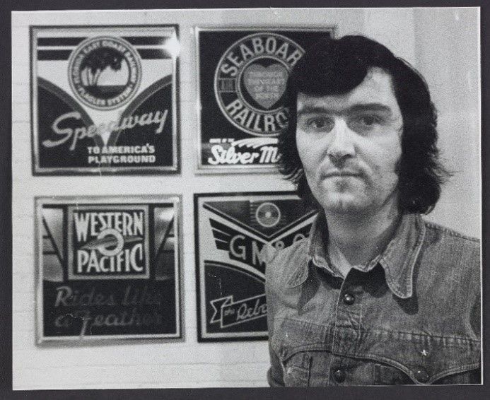 Black and white photograph of Ian Logan standing beside four American railroad logos he designed in the 1970s.