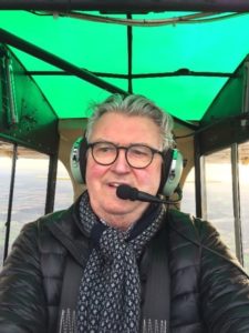 In this colour selfie, taken on his phone while flying his Piper Super Cub light aircraft, Ian Logan wears spectacles, headphones and microphone, patterned black-and-white scarf and black flying jacket.  