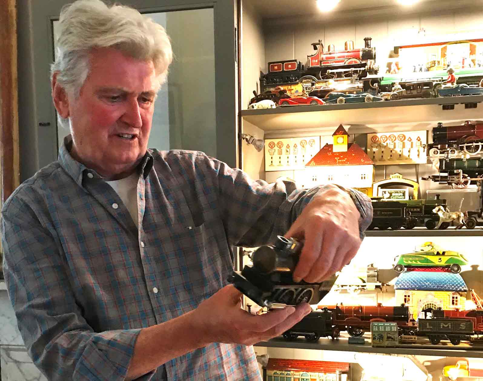 In this color photograph, the designer Ian Logan holds a model steam locomotive with an illuminated cabinet behind him containing other models.