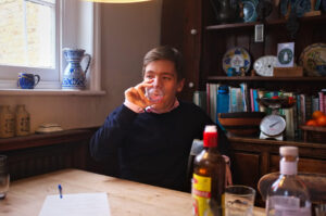 A man drinks from a small glass while sitting at a wooden table with bottles of cocktail ingredients on it.  