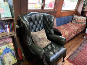 A dark green leather wing chair sits between the Children’s section and an ottoman piled with cushions.