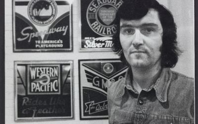 Black and white photograph of Ian Logan standing beside four American railroad logos he designed in the 1970s.