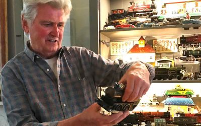 In this color photograph, the designer Ian Logan holds a model steam locomotive with an illuminated cabinet behind him containing other models.